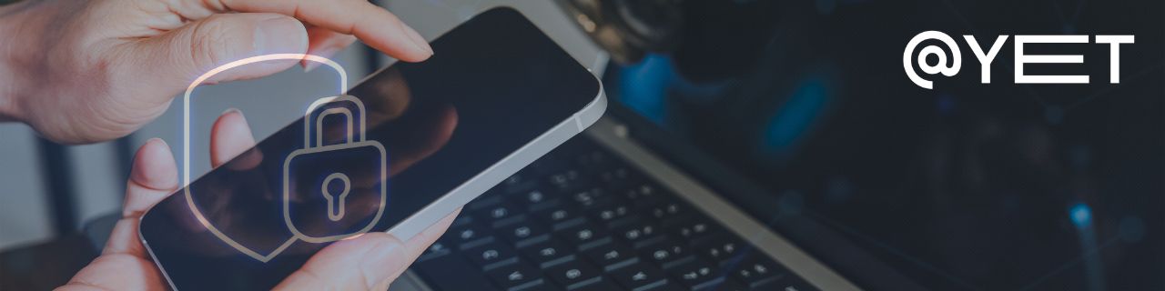 Im Hintergrund ein Laptop, im Vordergrund eine Hand, die ein Smartphine hält. Darüber transparent ein Schild mit einem Schloss als Symbol für Sicherheit